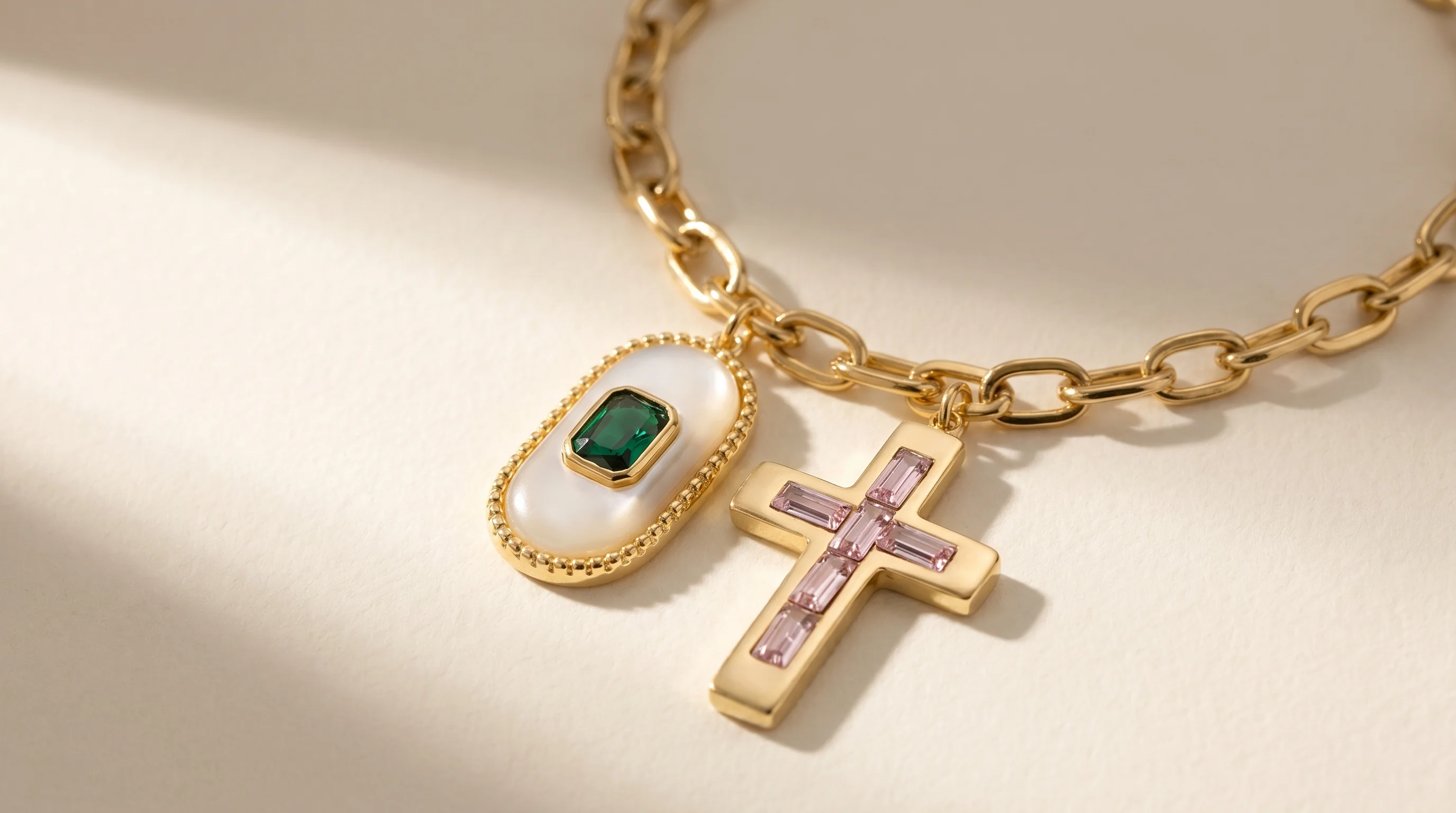 A gold link charm bracelet with a gemstone charm and a pink crystal cross charm on a cream background, by Enleira