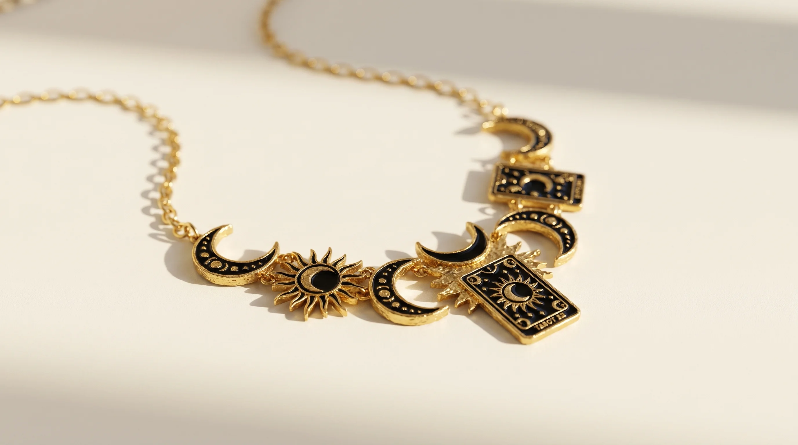 A gold celestial charm necklace with black enamel sun, moon, and tarot card motifs on a white surface, by Enleira