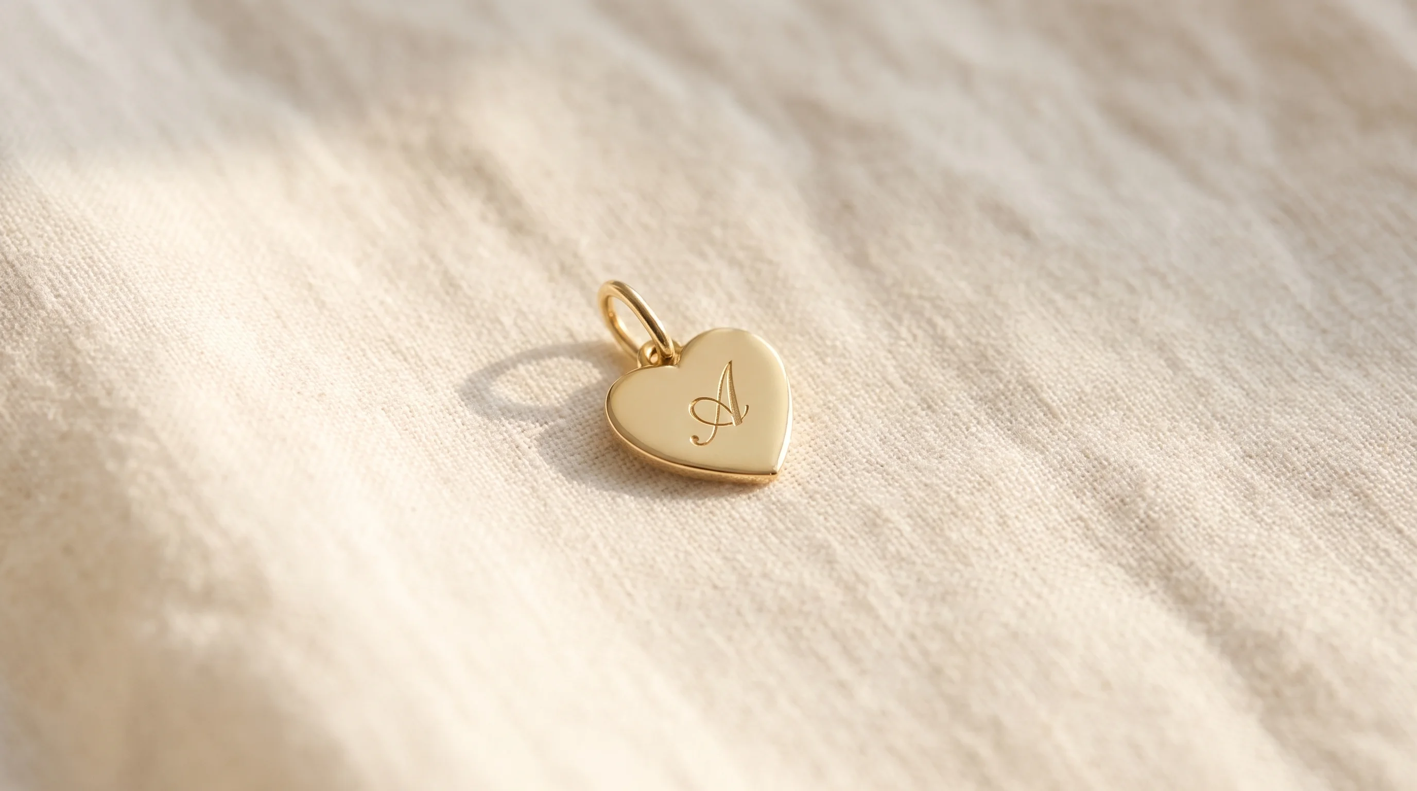 A gold heart-shaped charm engraved with the initial 'A' rests on a soft ivory linen surface in warm light, by Enleira.