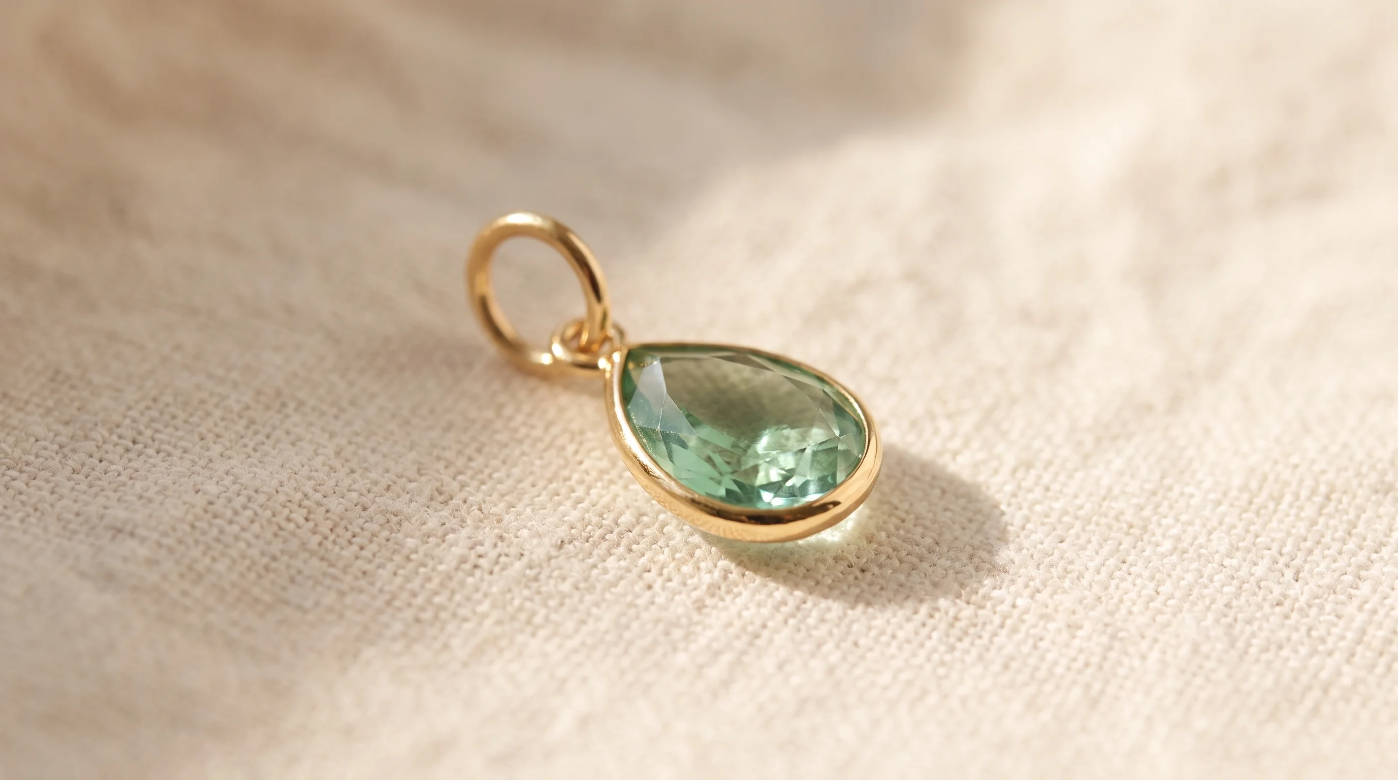 A gold charm with a faceted, teardrop-shaped green gemstone, resting on a cream-colored textured fabric, by Enleira