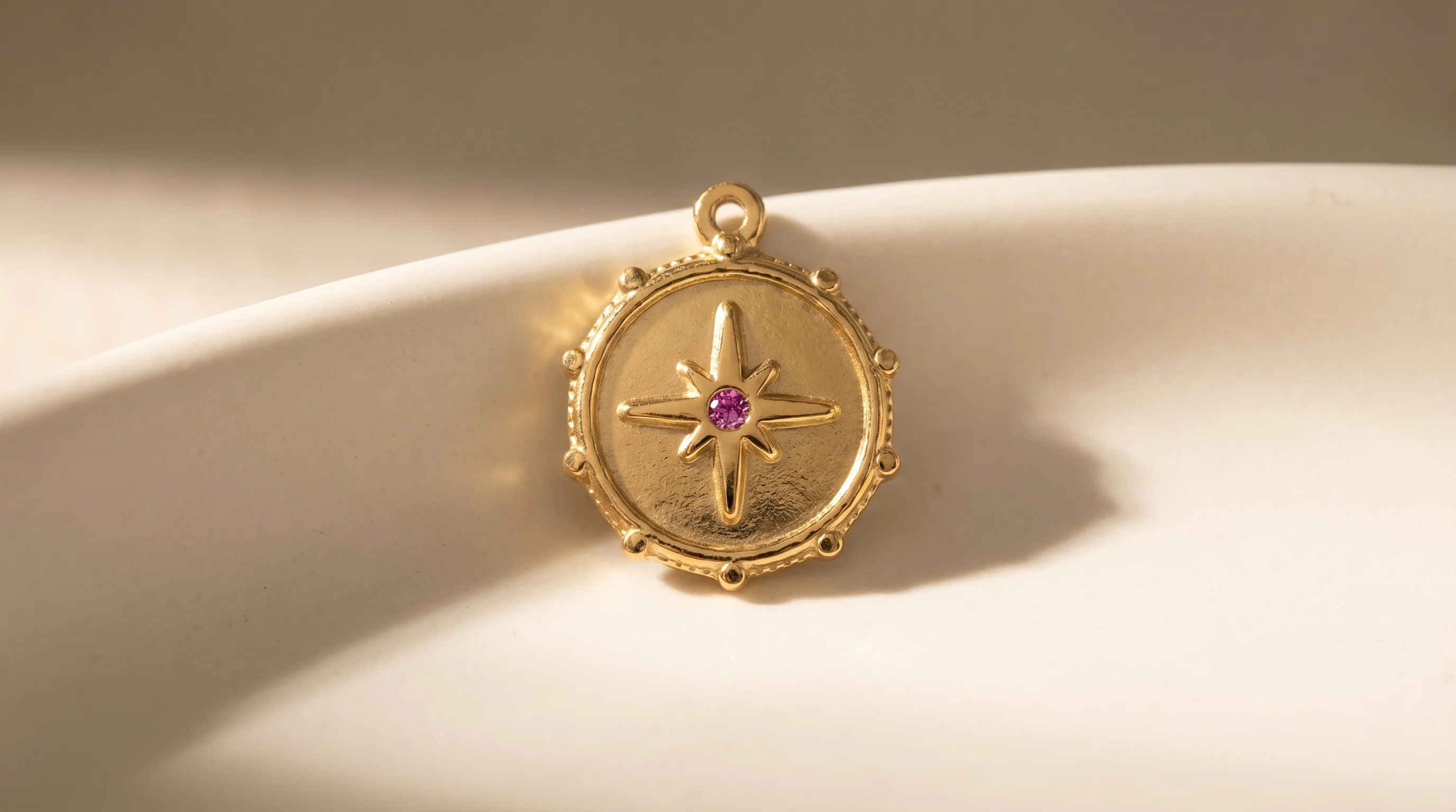 A handcrafted gold compass rose charm with a central pink gemstone, resting on a curved cream surface, by Enleira