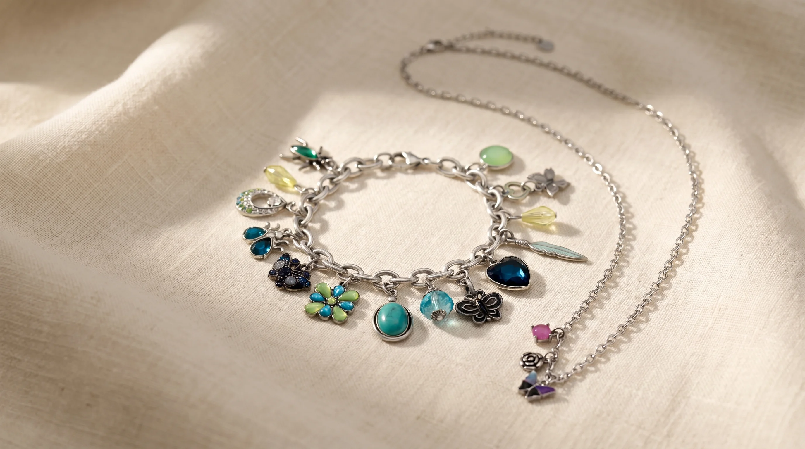 A silver charm bracelet and necklace with colorful gemstone charms arranged on a soft, cream-colored linen cloth, by Enleira